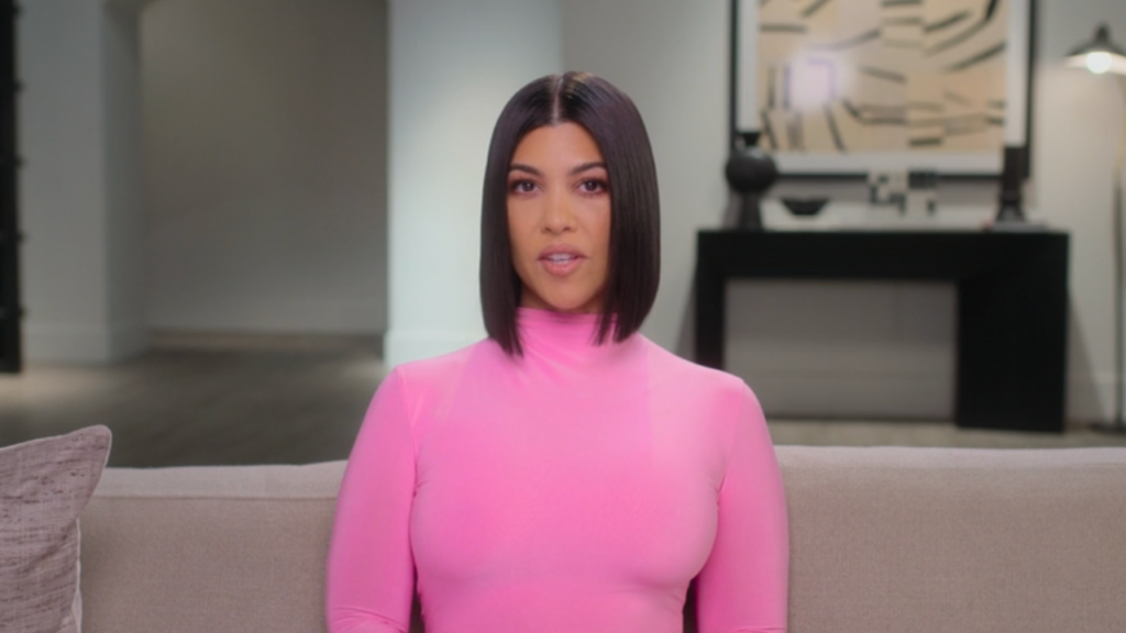 Kourtney Kardashian sits down smiling wearing a hot pink dress