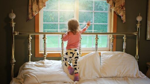 Little girl stands on a bed and points out a bedroom window