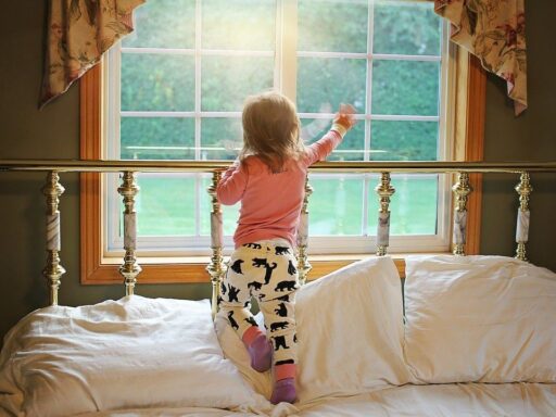 Little girl stands on a bed and points out a bedroom window