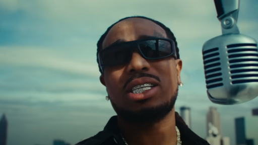 Quavo stands next to a microphone with sunglasses on and a city skyline behind him