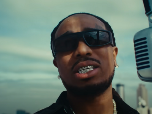 Quavo stands next to a microphone with sunglasses on and a city skyline behind him