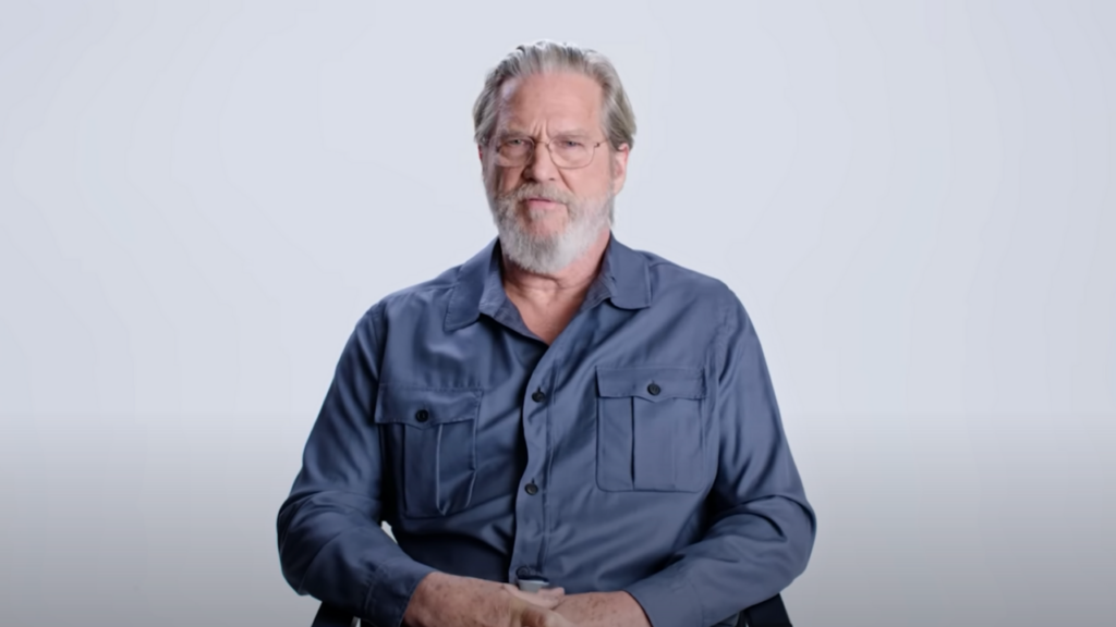 Jeff Bridges sits on a chair with blue shirt on with a white background