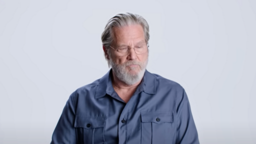 Jeff Bridges sits on a chair with blue shirt on with a white background