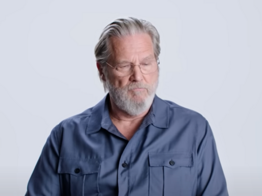 Jeff Bridges sits on a chair with blue shirt on with a white background