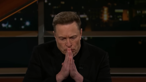 Elon Musk wears a black tee and blazer and sits in a chair