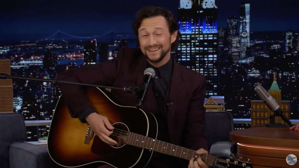 Joseph Gordon-Levitt wears a dark red suit, holds an acoustic guitar as he performs Lover by Taylor Swift